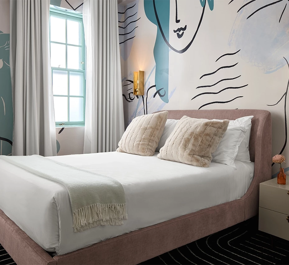 A neatly made bed with a pink upholstered frame and a white comforter, with two beige pillows and a throw blanket, in a room with a window, abstract wall art, and a nightstand.