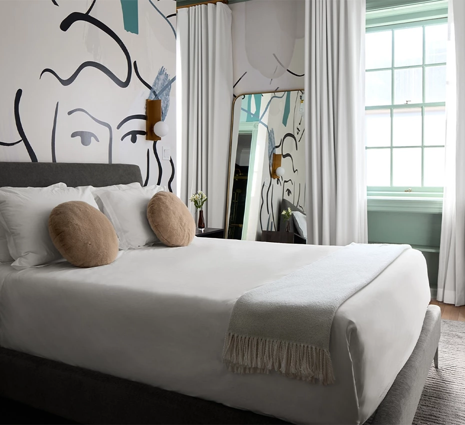 A neatly made bed in a room with a large window, abstract artwork on the wall, and a mirror.