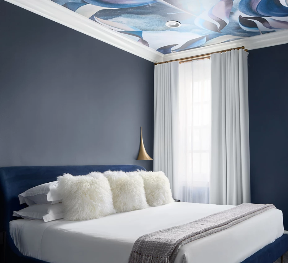 A luxurious hotel room with a blue accent wall, white bedding, and a mural on the ceiling.