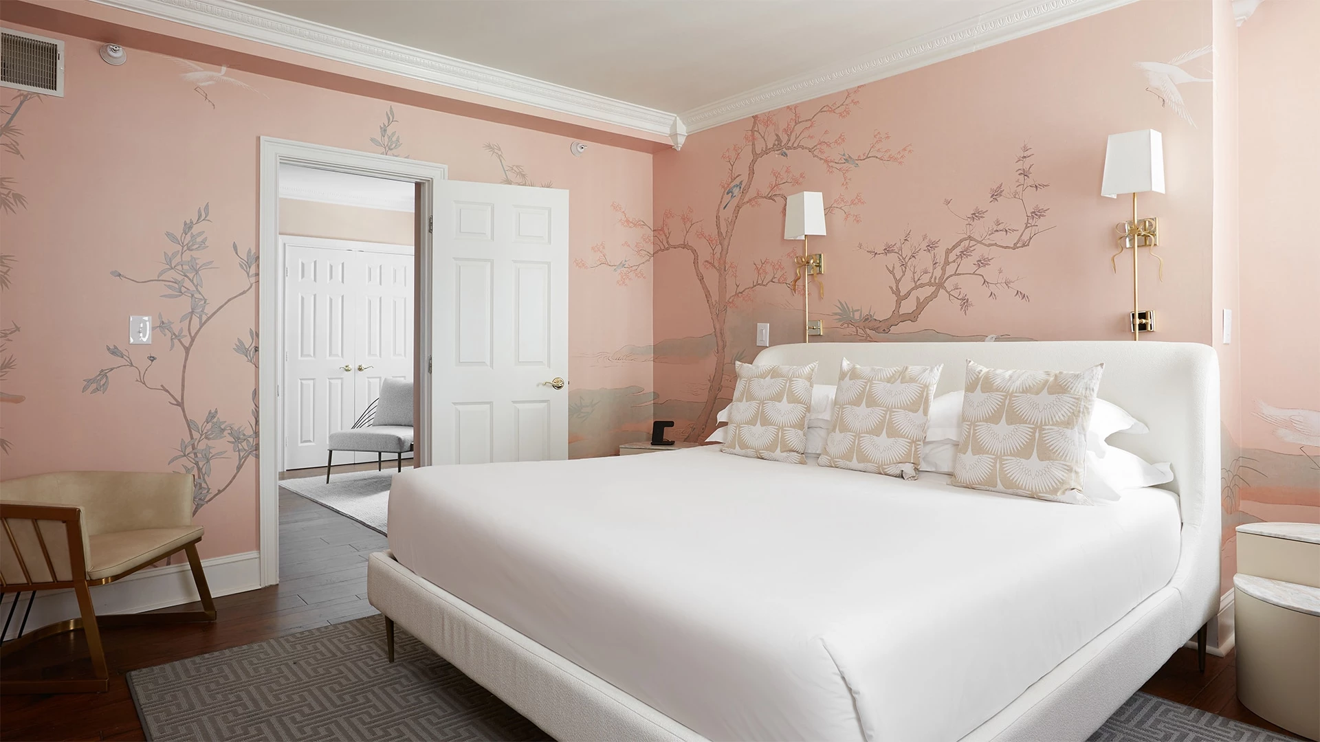 A wide view of a hotel room with a pink wall, a white bed, and a gold lamp.