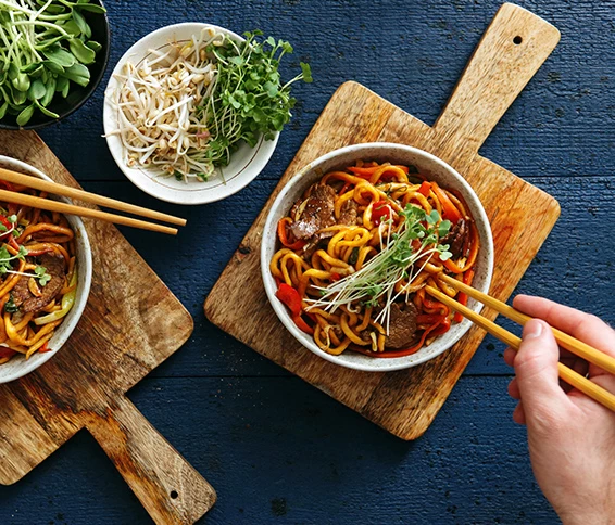 a top-down view of two bowls of noodles with vegetables and microgreens, served with chopsticks on wooden boards