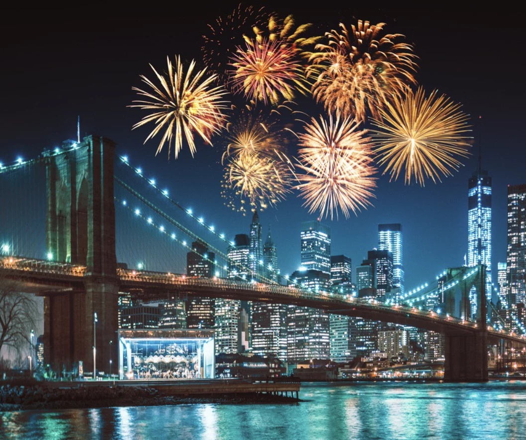 A nighttime view of the Brooklyn Bridge with fireworks exploding above it.
