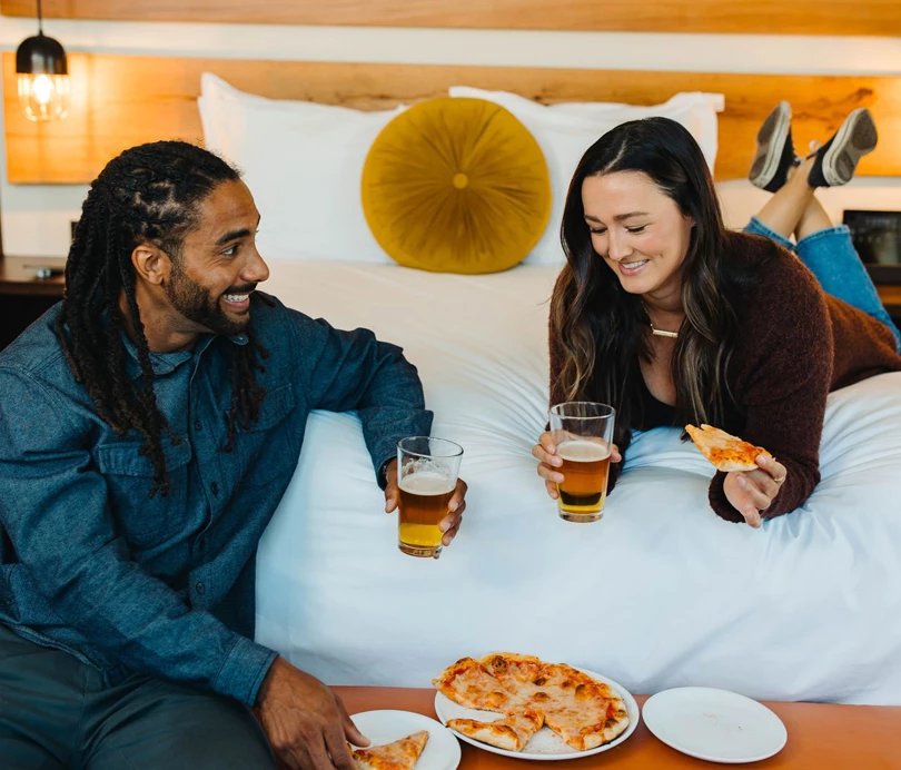 a man and woman sitting on a bed with pizza and beer