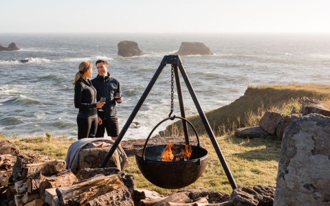 a man and woman standing next to a fire pit with a body of water in the background
