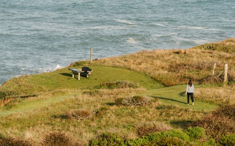 a woman standing on a grassy hill by a bench and water
