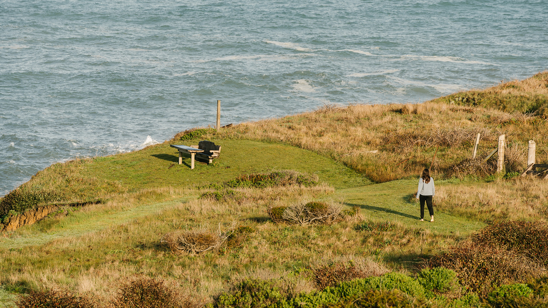 a woman standing on a grassy hill by a bench and water