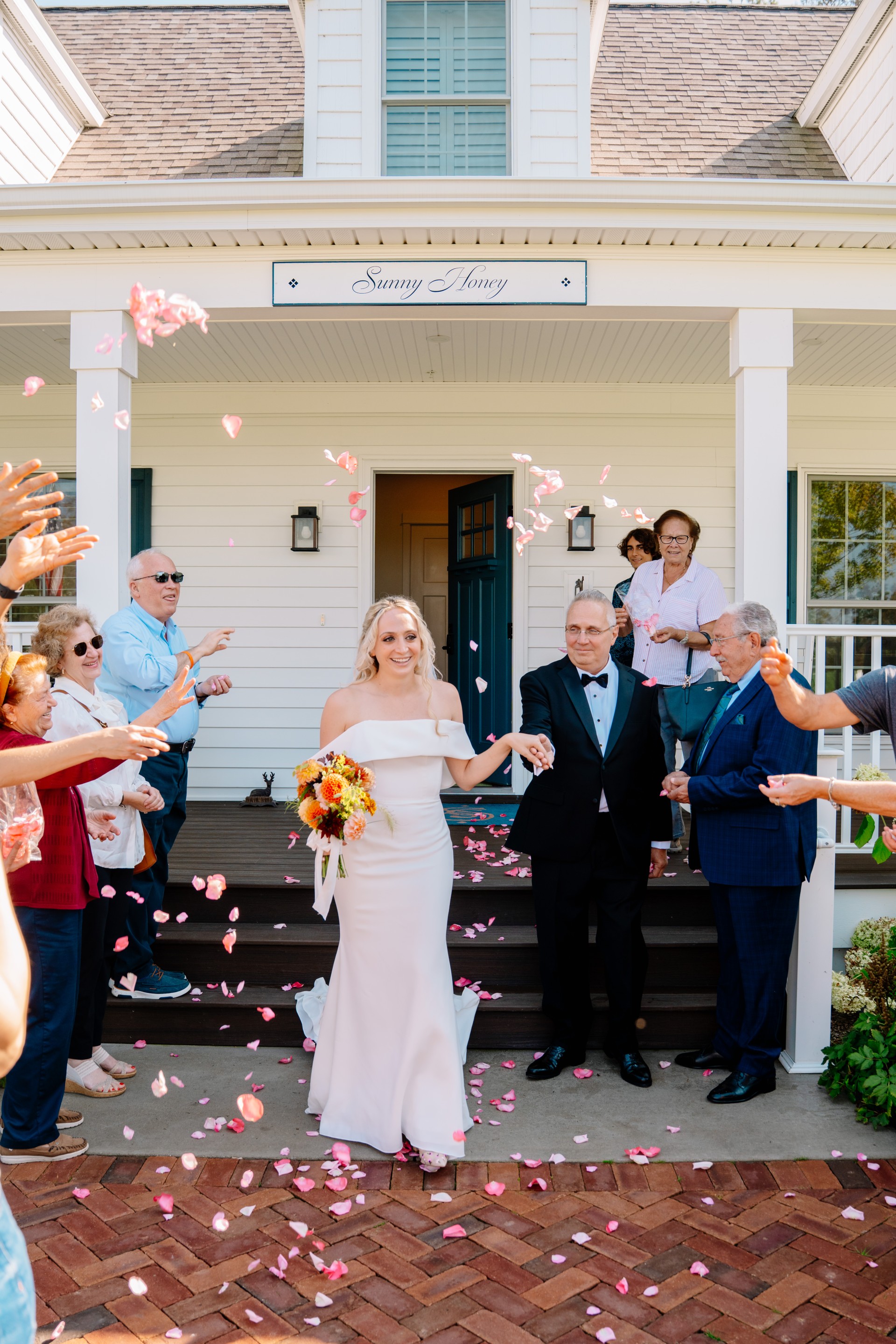 a woman in a white dress outside of a house with people throwing petals