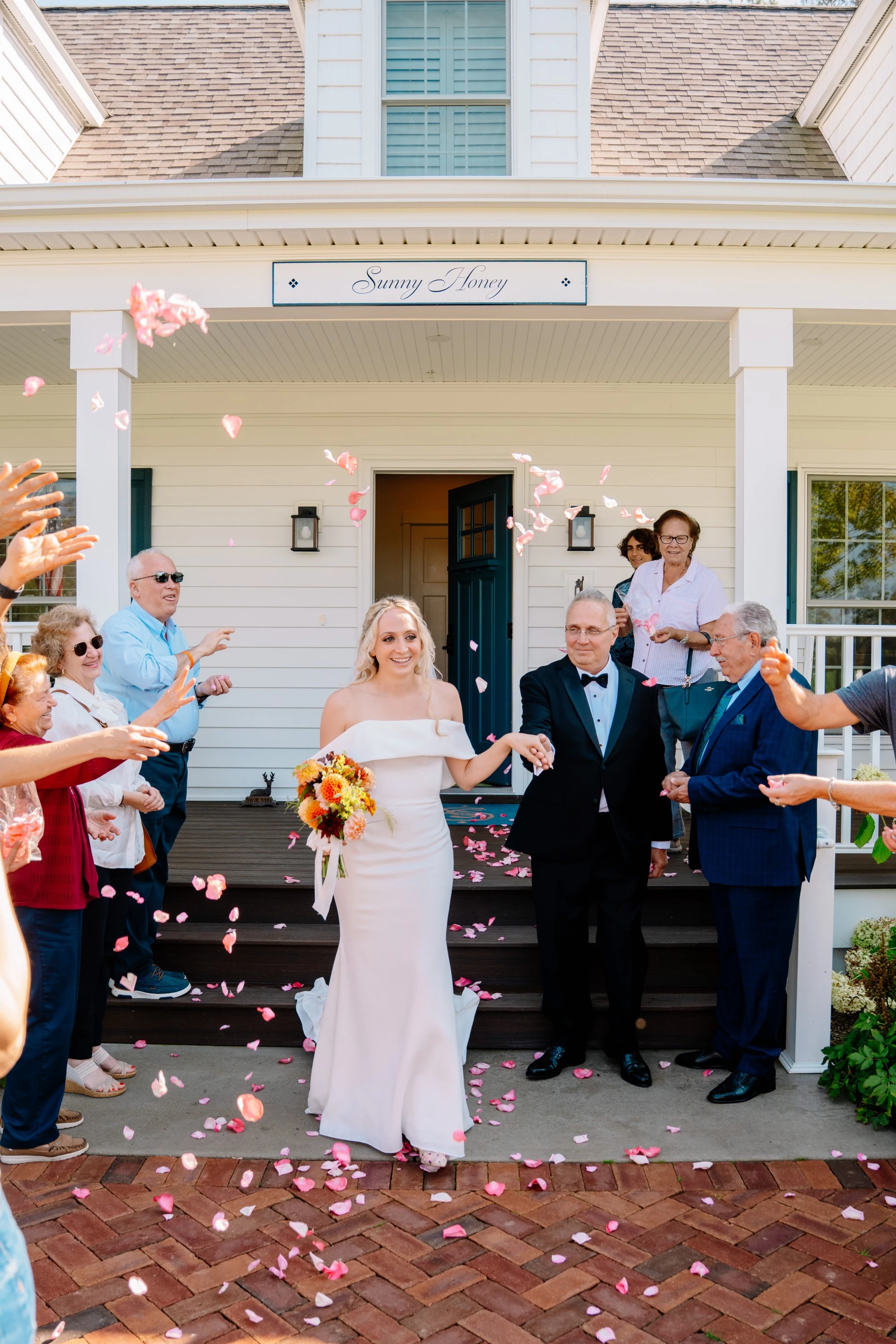 a woman in a white dress outside of a house with people throwing petals