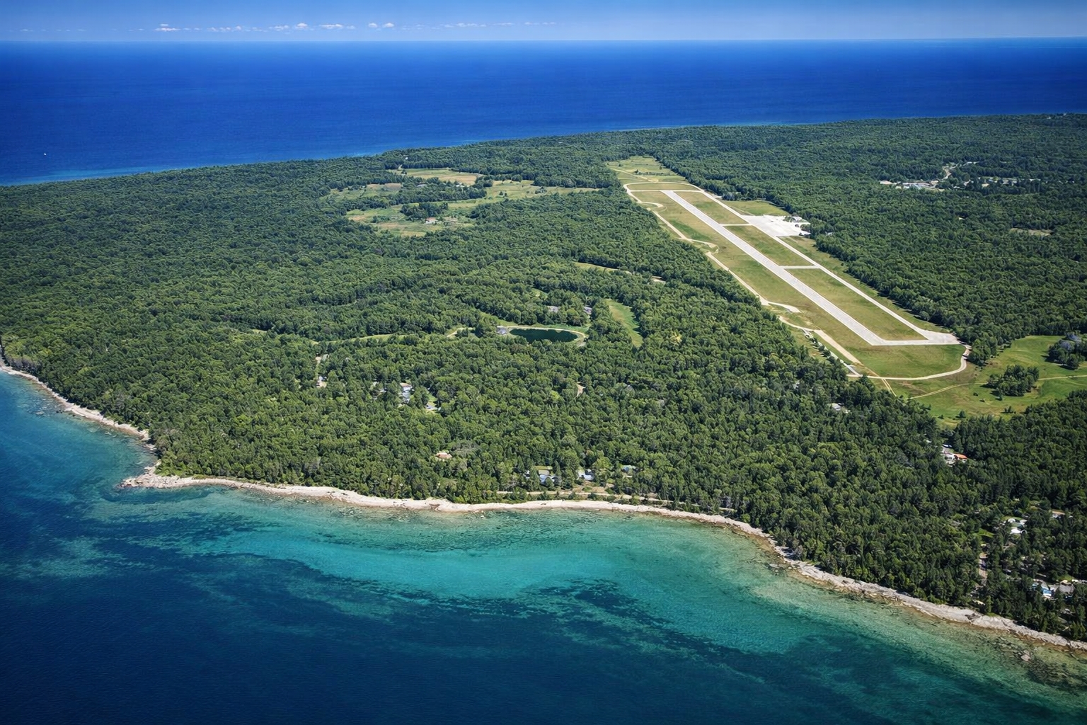 Aerial view of Mackinac Island Airport MCD with a small private plane landing, surrounded by Lake Huron, forest, and proximity to The Inn at Stonecliffe in spring