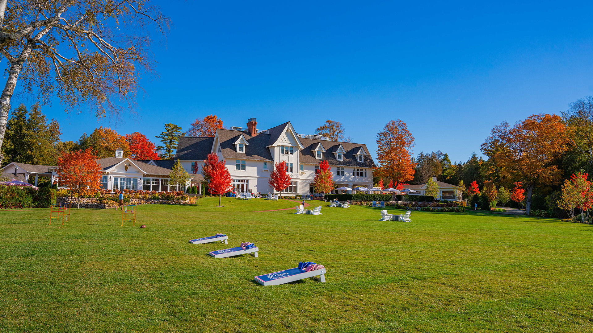 Expansive green lawn with cornhole games and white chairs in front of The Inn at Stonecliffe, a historic mansion-style hotel on Mackinac Island surrounded by vibrant fall foliage