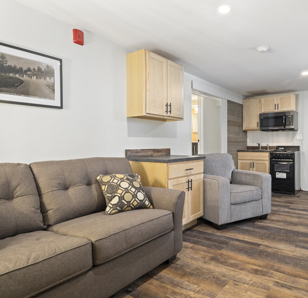 2 bedroom apartment room shot of a small pantry and kitchen with an oven, microwave, sink, a two person couch and a single lounge chair in the living room area