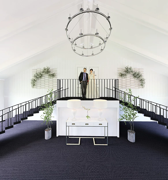 a man and woman standing on a white table in a room with stairs and a chandelier