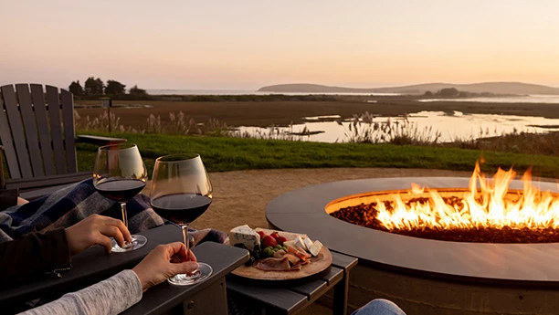 people holding wine glasses and sitting at a table with food and a fire pit