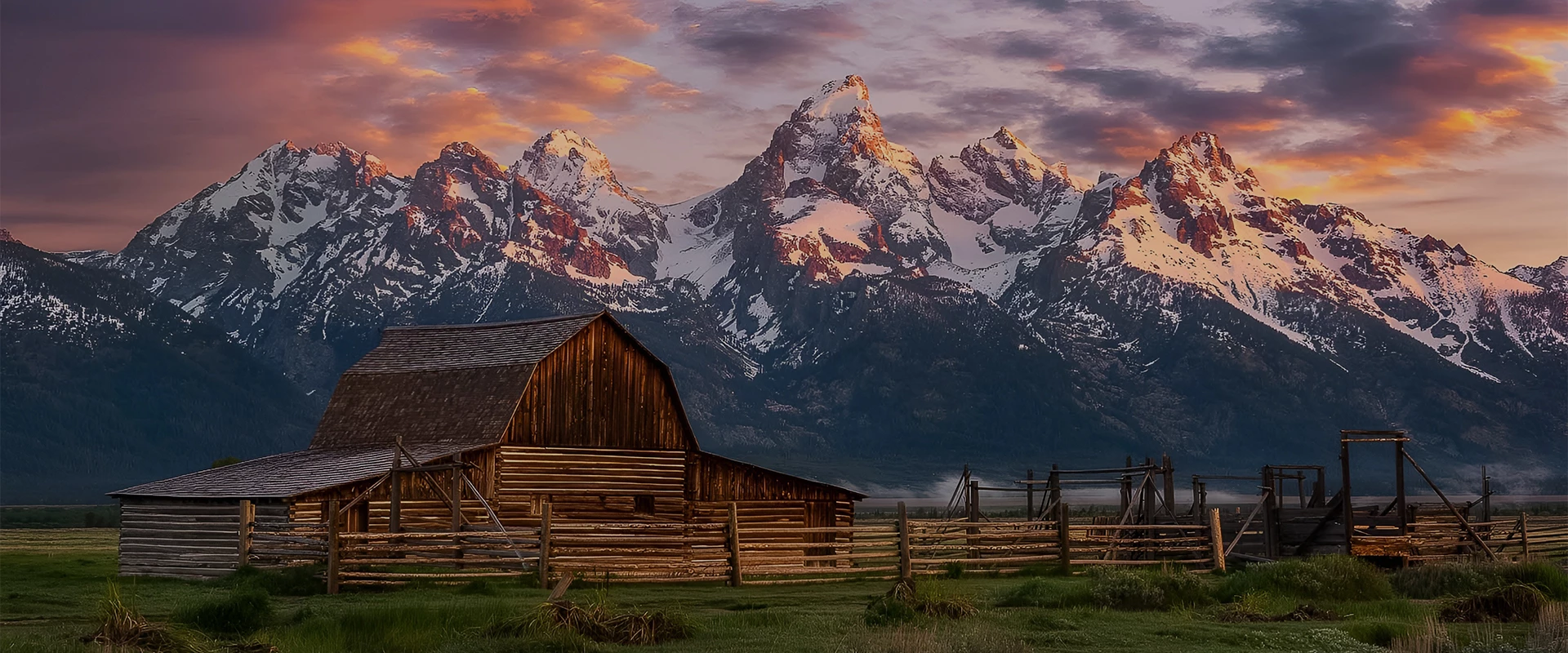 a barn in a field with snow covered mountains in the background