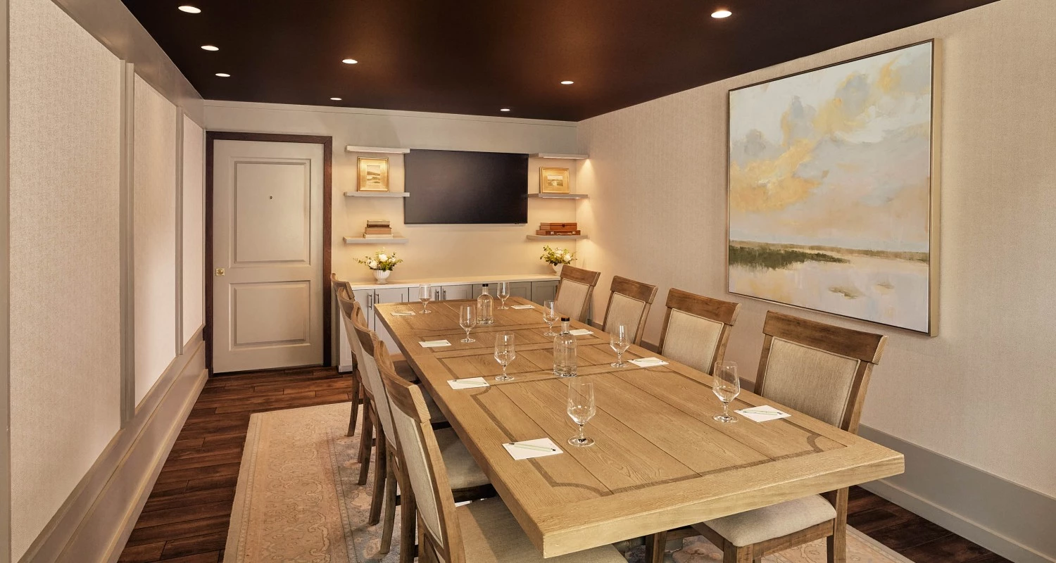A meeting room with a long wooden table and chairs, a painting on the wall, and a TV mounted on the wall.