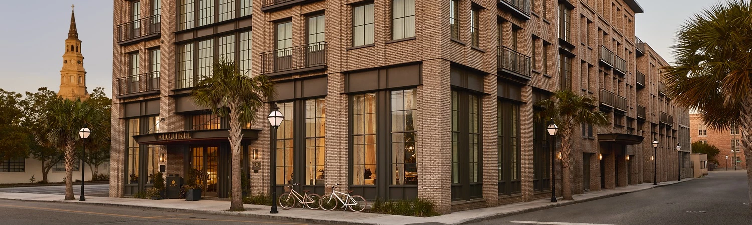 A multi-story brick building with balconies and large windows, situated on a street corner with palm trees and bicycles outside.