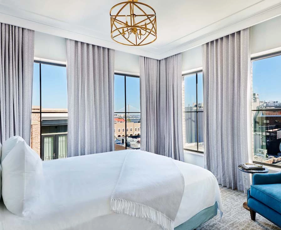 A wide view of a hotel room with a large bed, blue chair, and city view.