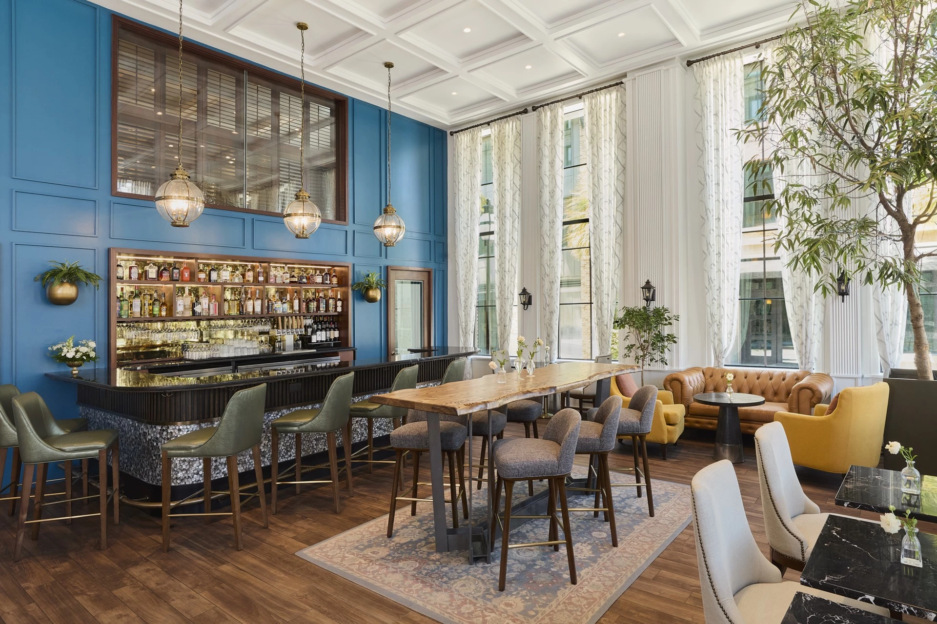 A stylish bar area with a blue accent wall, a bar with stools, a large table with chairs, and comfortable seating areas.