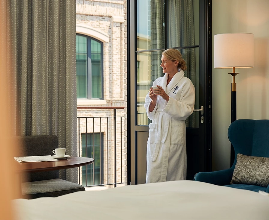 A woman in a white robe stands by a balcony door with a city view, next to a bed and chair.
