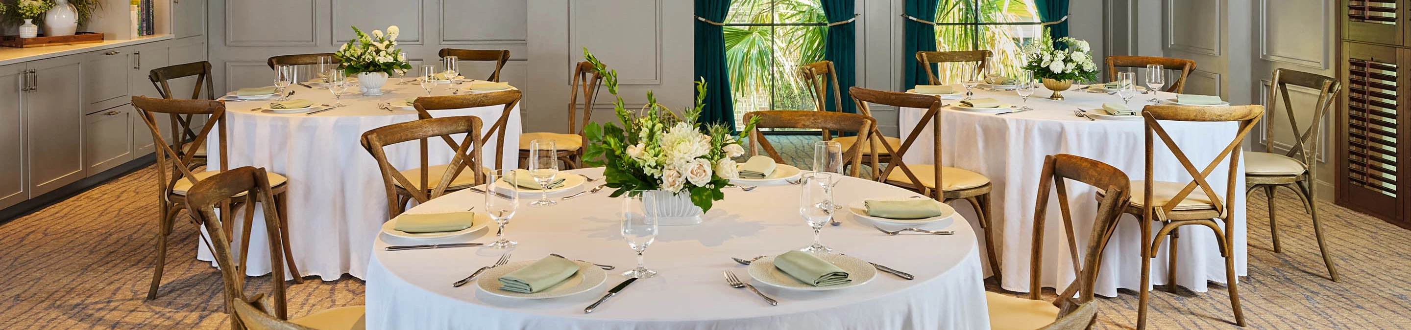 A dining room with round tables set for a meal, featuring white tablecloths, wooden chairs, and flower centerpieces.