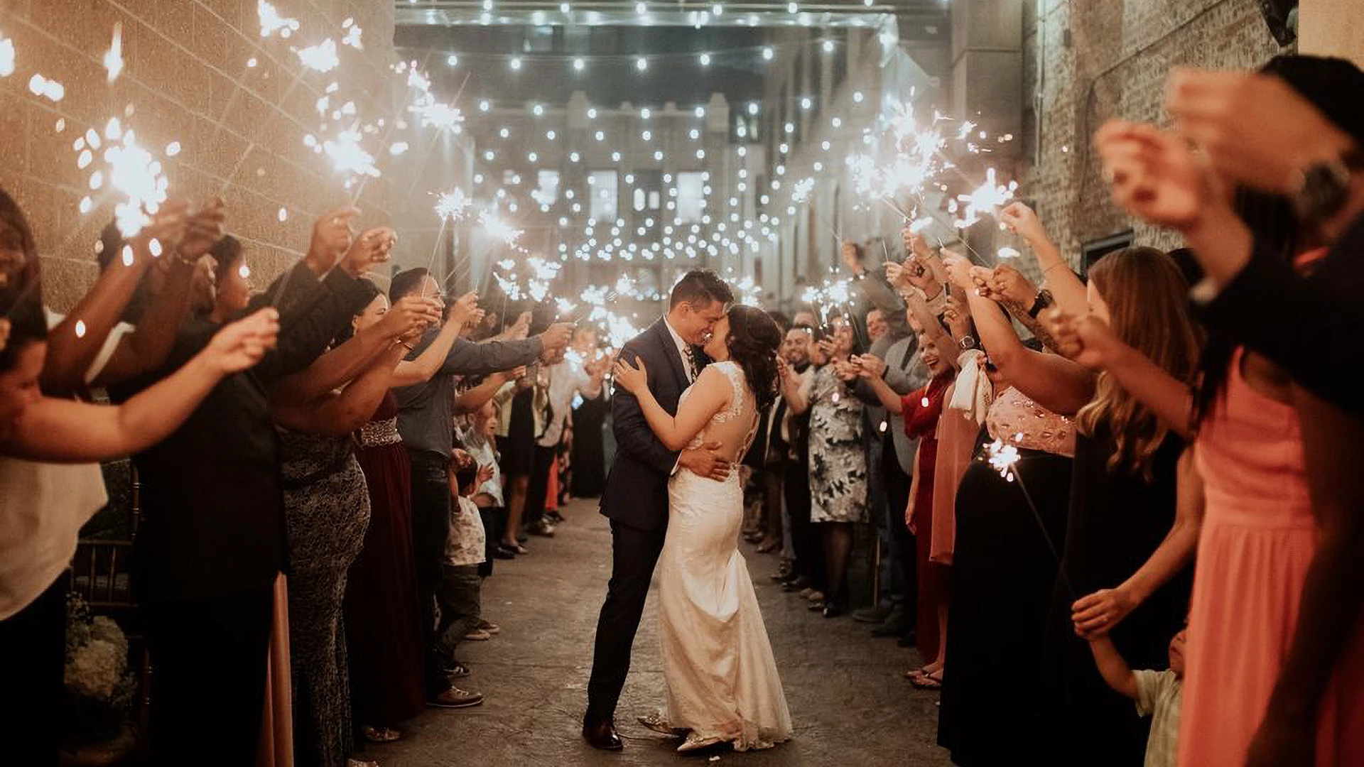 A couple kisses in a corridor with people holding sparklers.