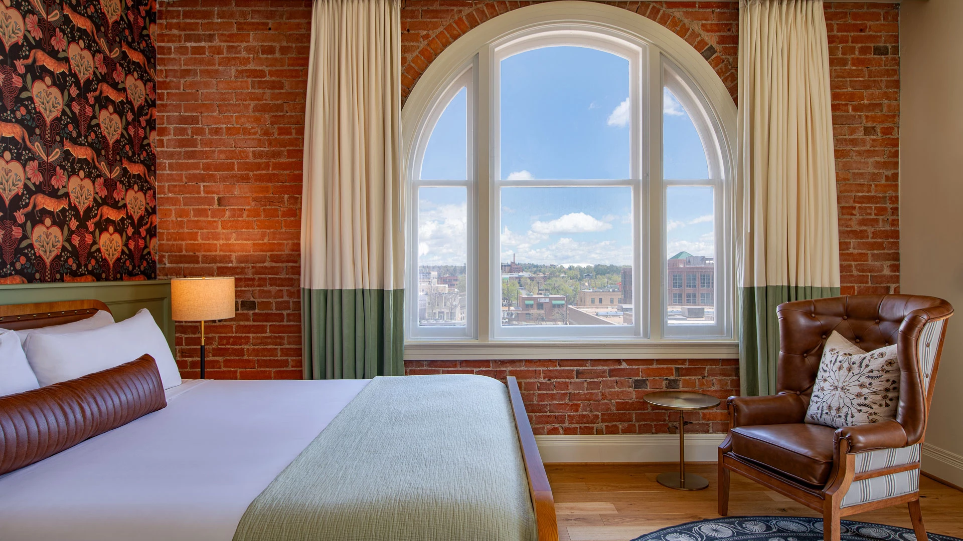 A hotel room with a bed, chair, and window overlooking the city.