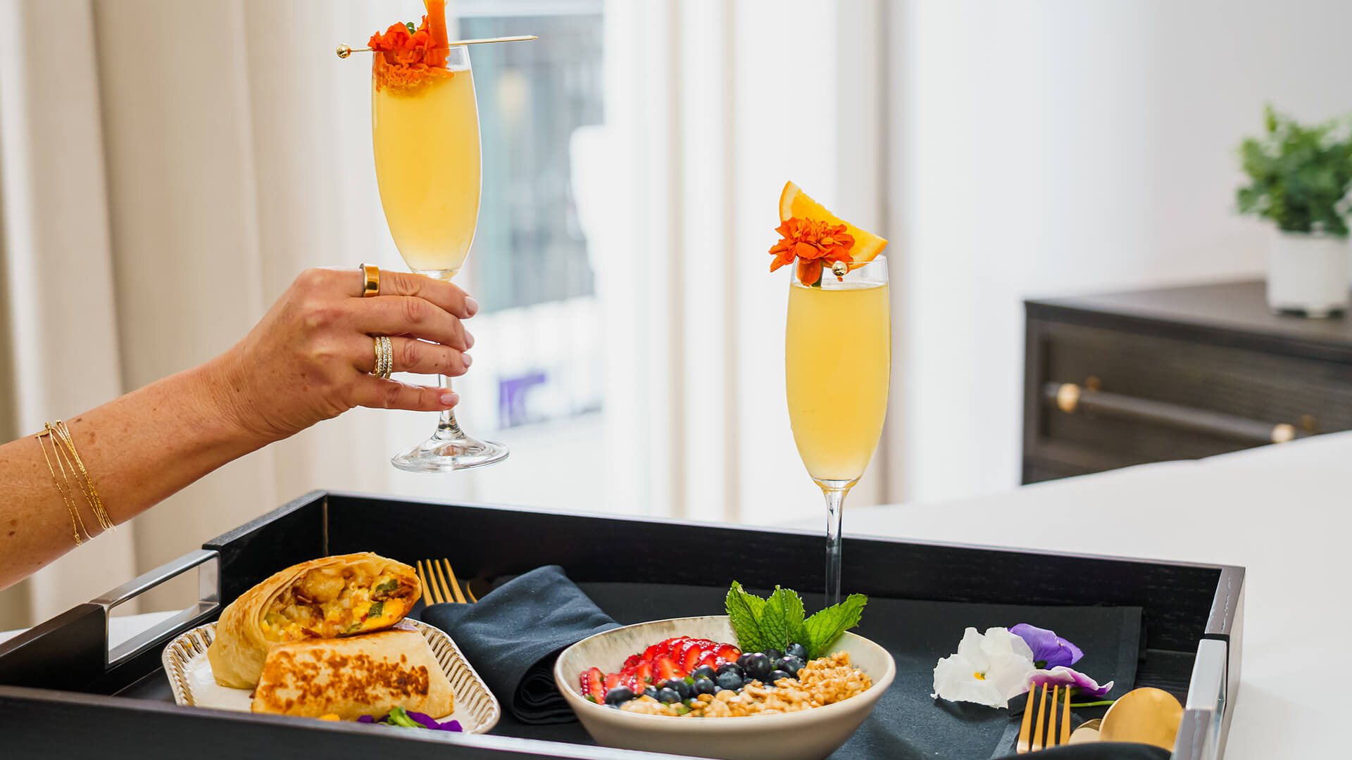 a hand holding a glass of yellow liquid with orange flowers and a plate of food