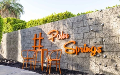 A stone wall with orange lettering reading 'Palm Springs' and three orange chairs in front of it.
