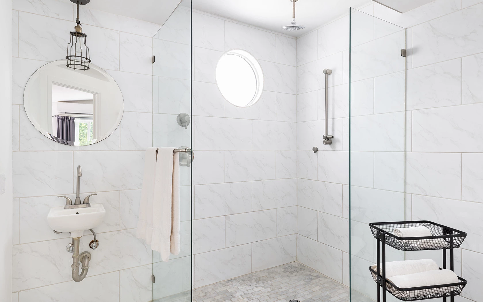 A modern bathroom with a glass-enclosed shower, marble tiles, and a round mirror.