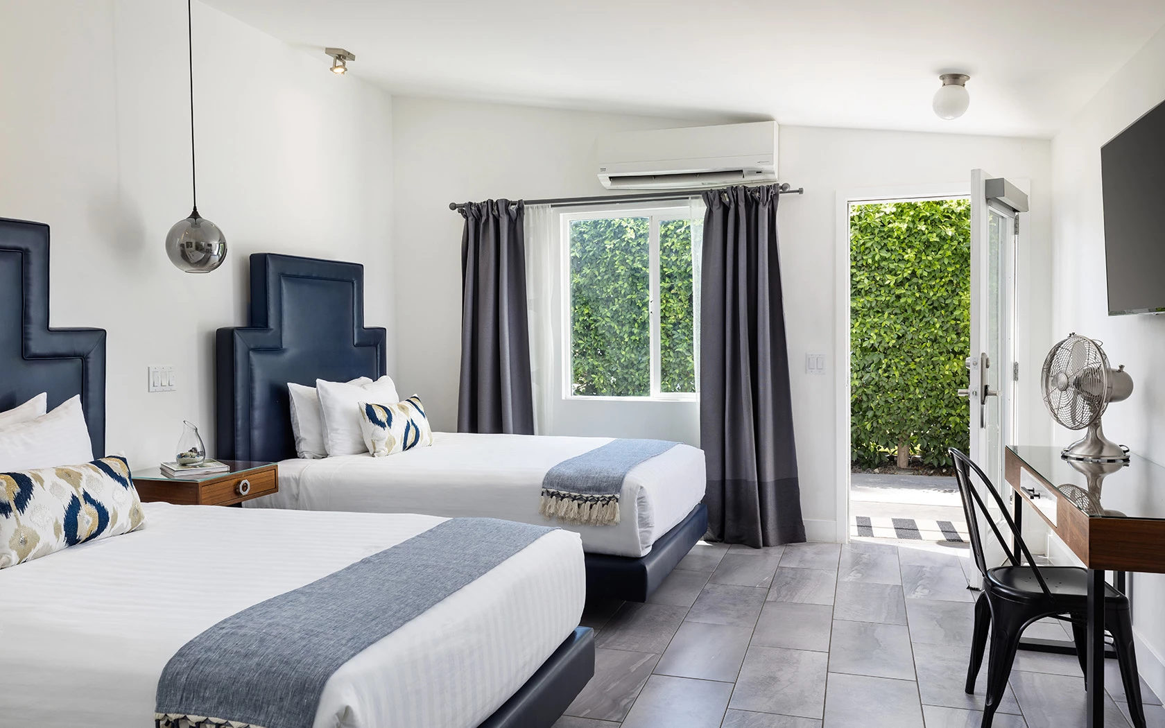 A hotel room with two beds, a window with a view of greenery, and a desk area with a chair and TV.
