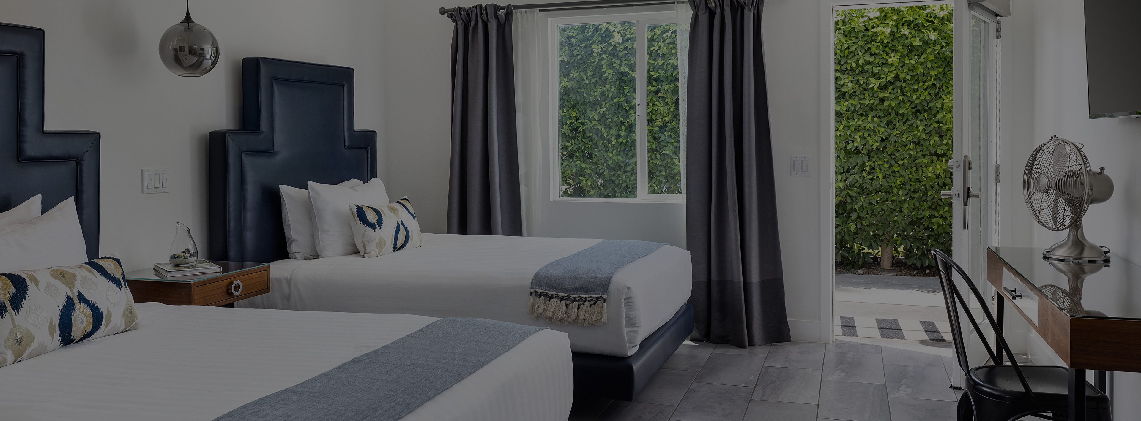 A hotel room with two beds, a desk, a window with curtains, and an open door
