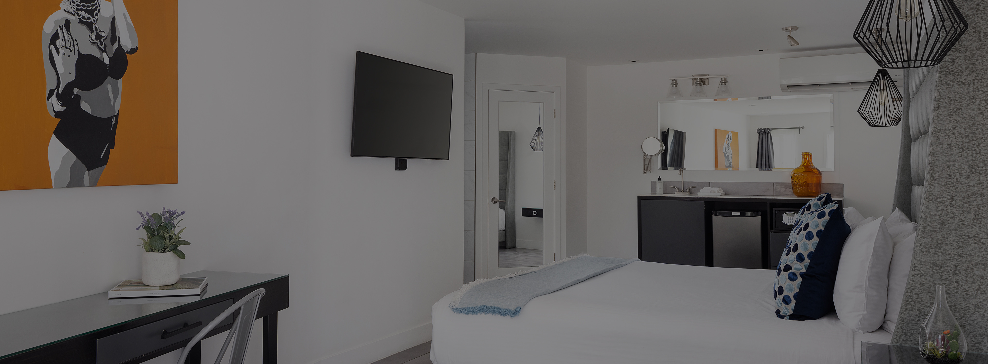 A modern hotel room with a bed, TV, desk, and kitchenette.
