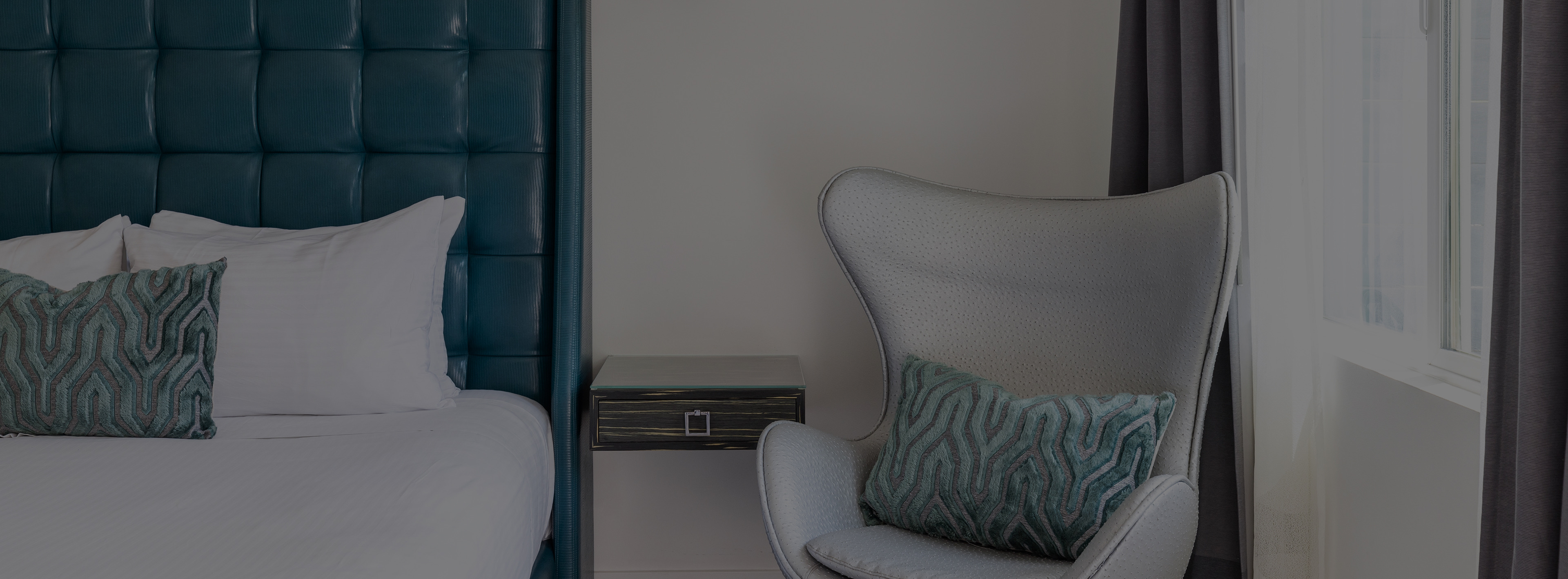 A bed with a tufted teal headboard and white linens, accompanied by a wingback chair and nightstand.