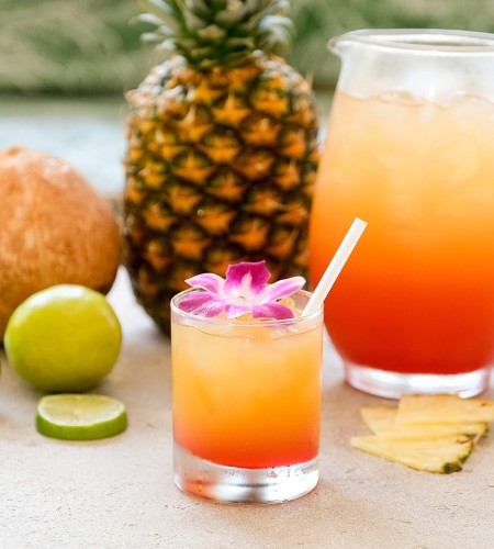 a glass of orange and yellow drink next to a pitcher of ice and fruit