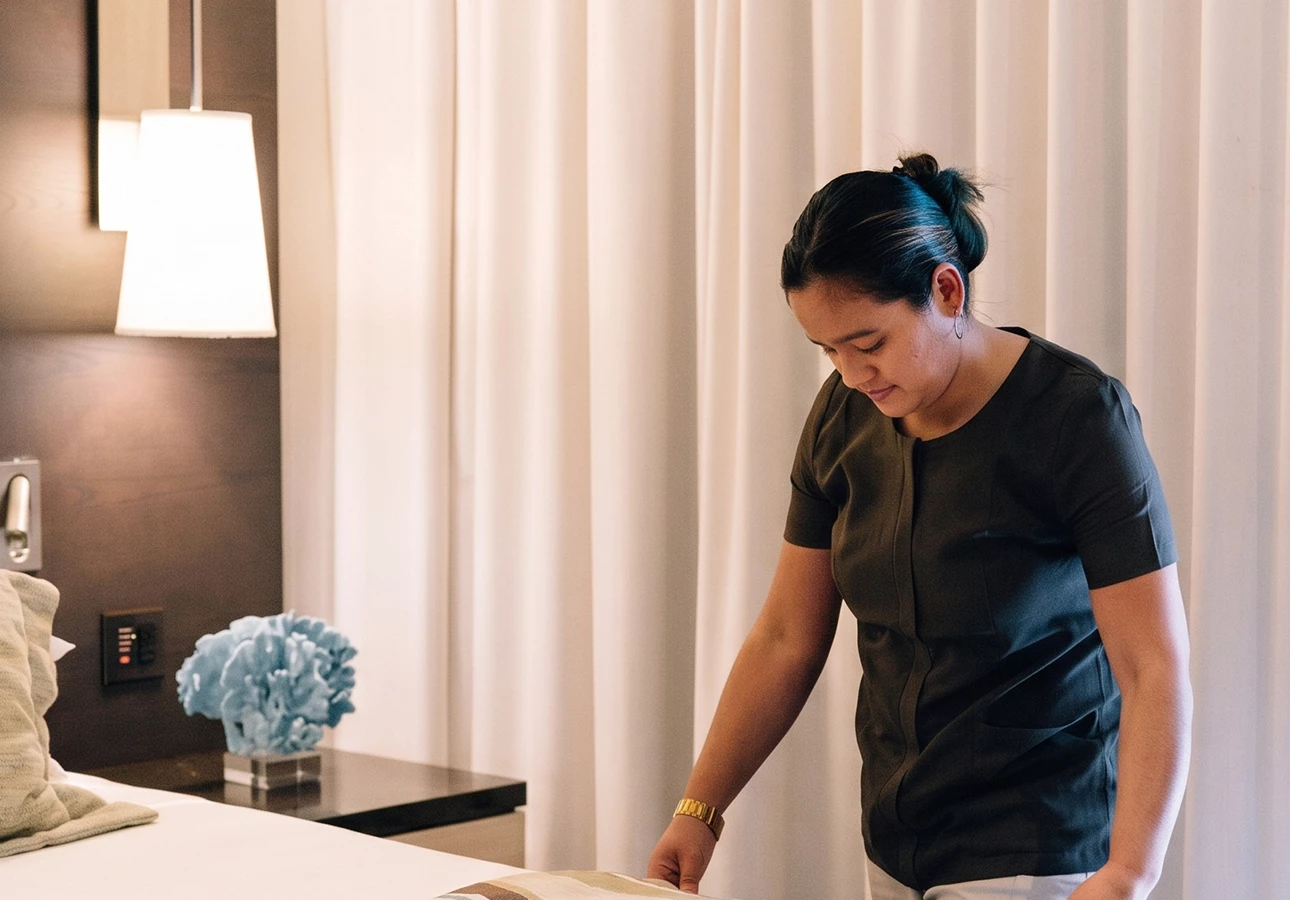 A hotel room with a neatly made bed, a nightstand with a blue floral arrangement, and a staff member in a black uniform.