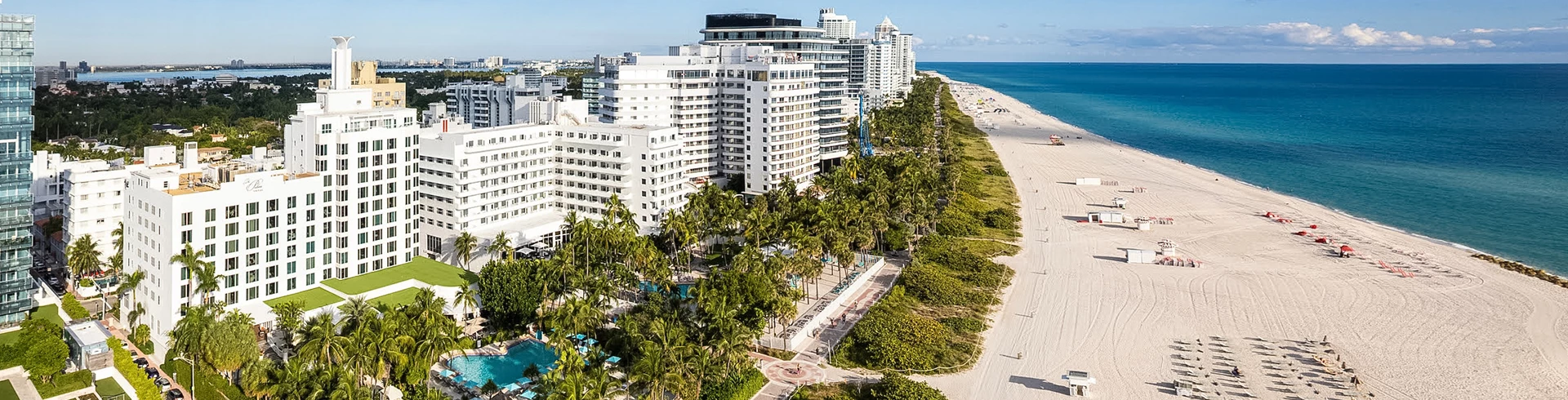 An aerial view of a white hotel building with a pool and palm trees on a beachfront property.