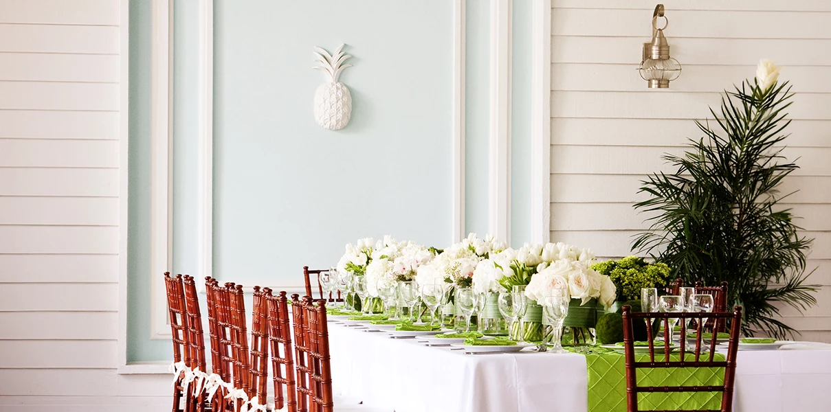 A formal dining area with a long table set with white flowers, green place mats, and red chairs.