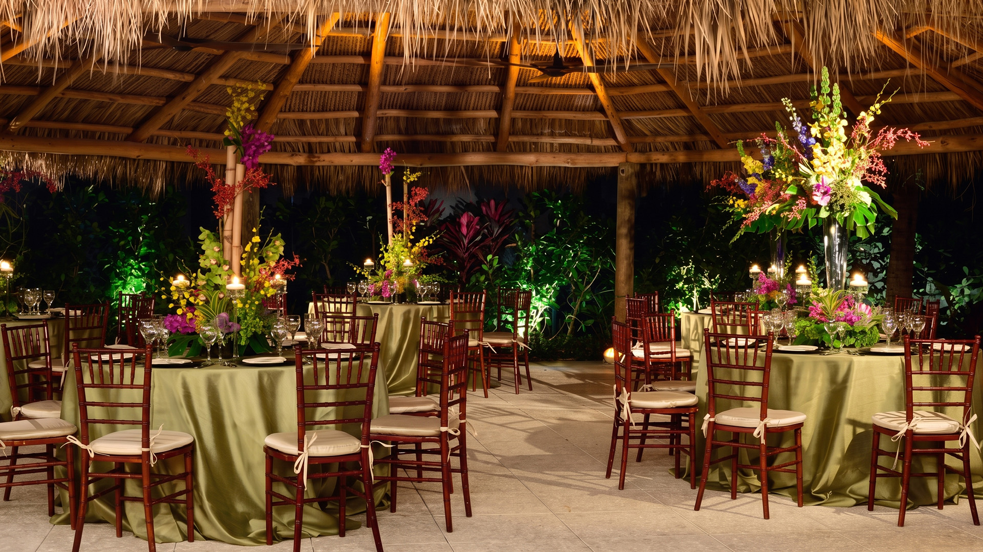 A dining area with round tables set for an event, featuring floral arrangements, green tablecloths, and thatched roof.
