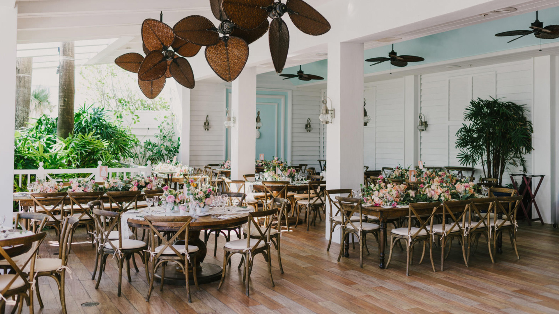 A large, brightly lit dining room with round tables set for an event, featuring floral arrangements, ceiling fans, and large windows.
