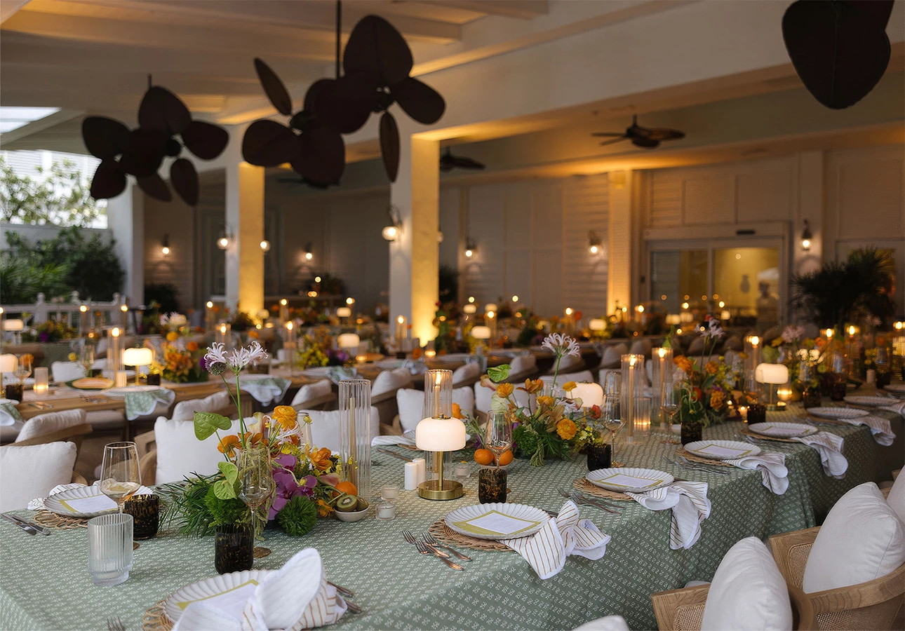 A beautifully set dining area with long tables, white chairs, green tablecloths, flower arrangements, and candle lighting.