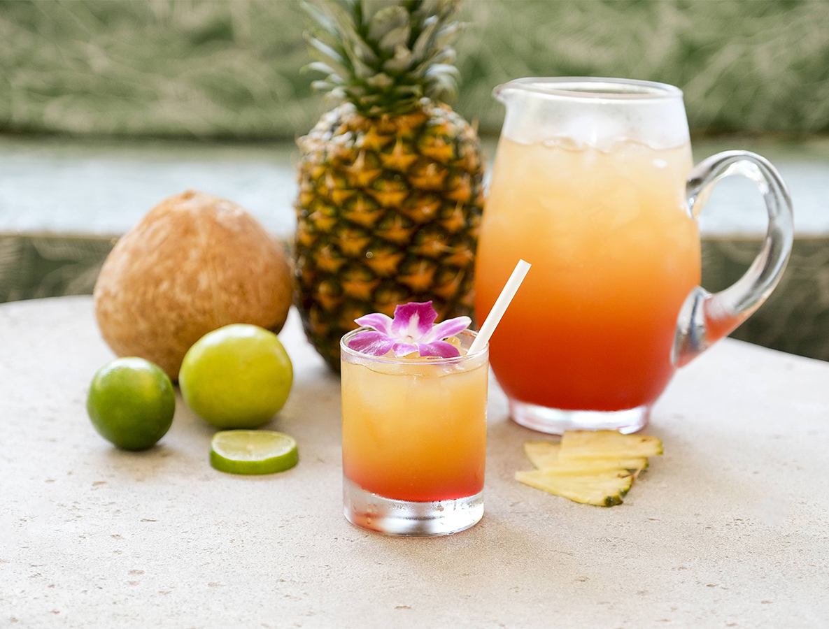 a glass pitcher and a drink with a straw next to a pineapple and limes