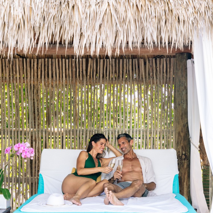 a man and woman sitting on a bed under a straw canopy