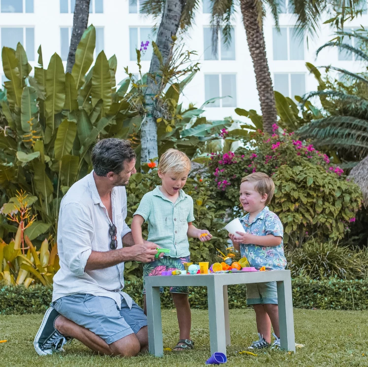 A man and two boys playing with toys on a table in a tropical garden with a large building in the background.