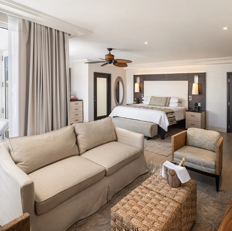 A wide view of a hotel suite with a beige sofa, wicker coffee table with champagne, and a large bed in the background.