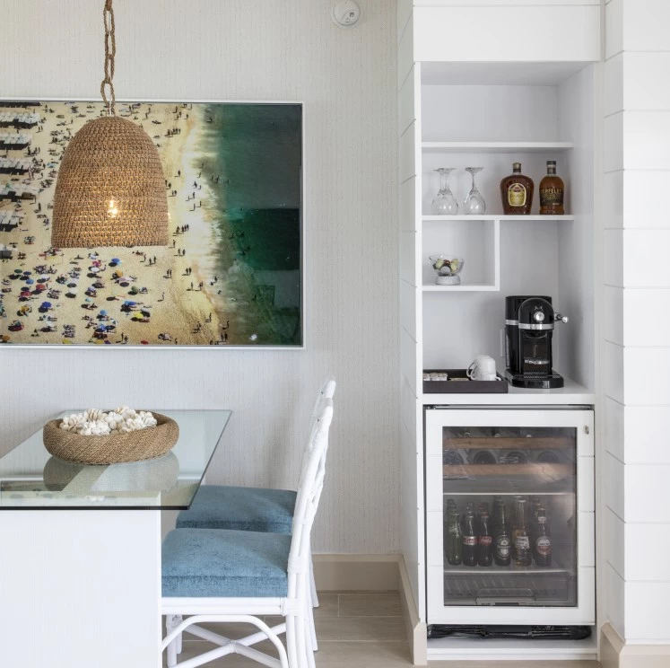 A compact kitchenette with a glass table, white cabinetry, mini fridge, coffee maker, and wine glasses.