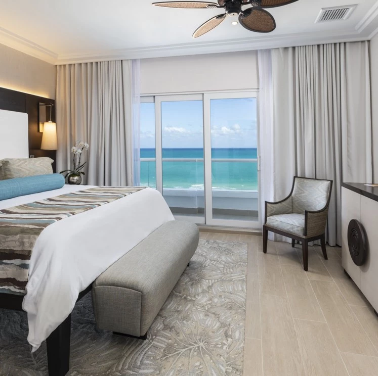 A wide-angle view of a hotel room with a large bed, comfortable seating, and a stunning ocean view.