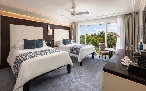 A wide view of a hotel room with two beds, a desk, and a window with a view of trees.