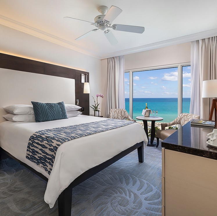 A wide-angle view of a hotel room with a large bed, ocean view, and modern furnishings.