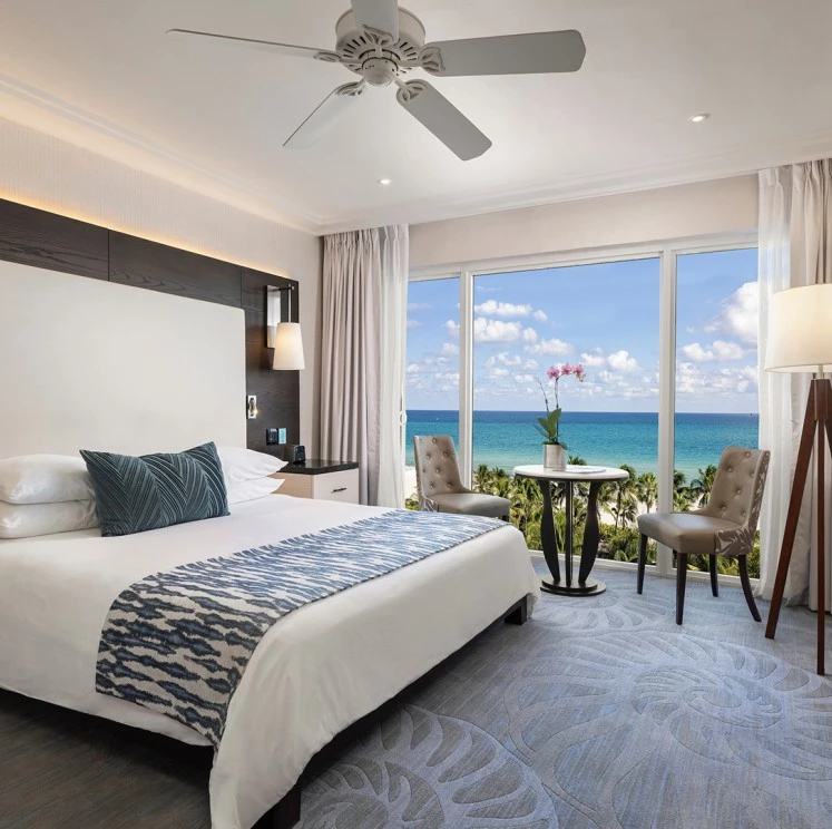 A wide shot of a hotel room with a large bed, two chairs, and a table in front of a large window with an ocean view.