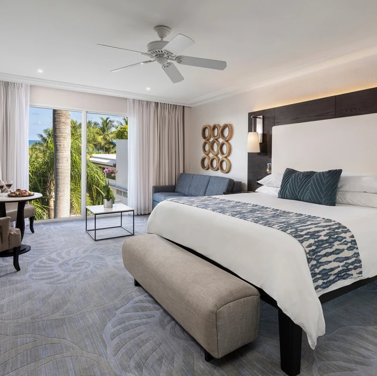 A wide view of a hotel room with a large bed, sofa, coffee table, and sliding glass doors leading to a balcony with palm trees outside.